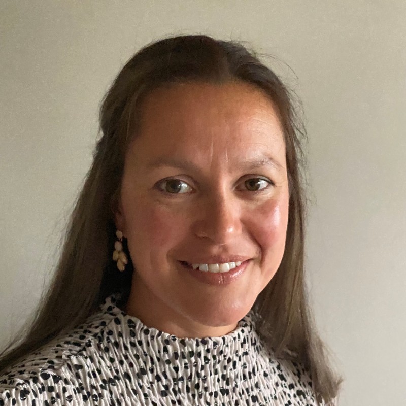 A headshot photo of Sarah Luke, a female with tanned skin and long, straight, brown hair. Sarah wears a cream and black spotted blouse.
