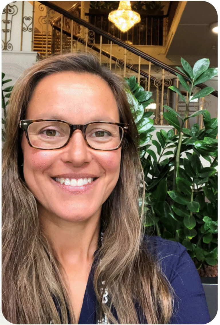 A headshot photo of Sarah Luke, a female with tanned skin, dark framed glasses, and long, straight, brown hair. Sarah wears a navy dress.