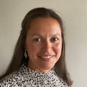 A headshot photo of Sarah Luke, a female with tanned skin and long, straight, brown hair. Sarah wears a cream and black spotted blouse.